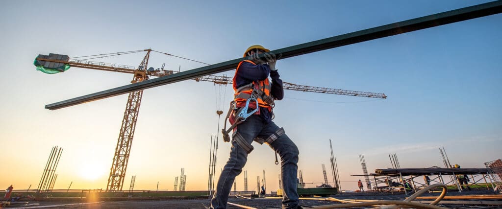 Ouvrier du bâtiment portant une poutre métallique sur un chantier au coucher du soleil, avec grue et équipement de sécurité.