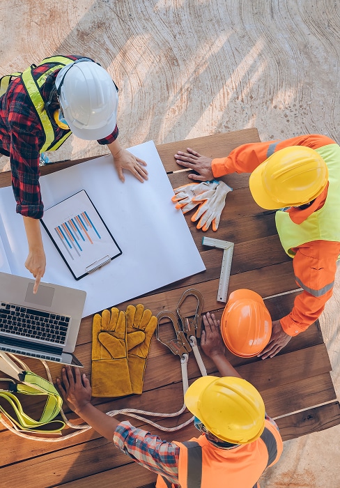 Trois ouvriers du bâtiment en casques et gilets regardent plans et laptop sur table en bois. Équipement de sécurité.