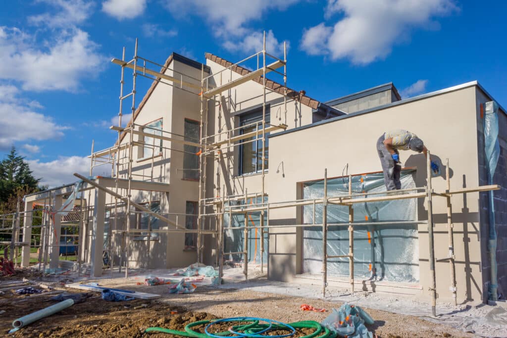Maison en construction. Un plâtrier applique l'enduit sur la façade beige d'une maison neuve avec échafaudage sous un ciel bleu.