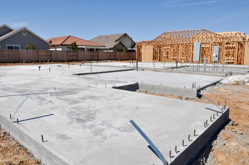 Nouvelle construction résidentielle. Dalle de fondation en béton au premier plan, charpente en bois d'une maison en arrière-plan sous un ciel bleu.