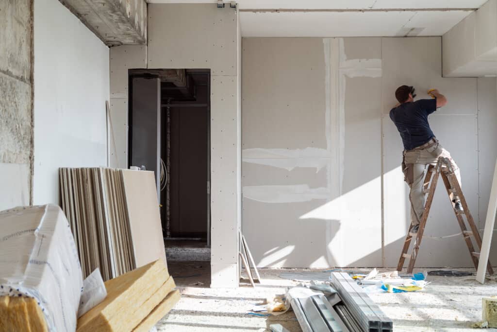 Ouvrier sur échelle posant des plaques de plâtre sur les murs d'un chantier de rénovation. Matériaux de construction au sol.