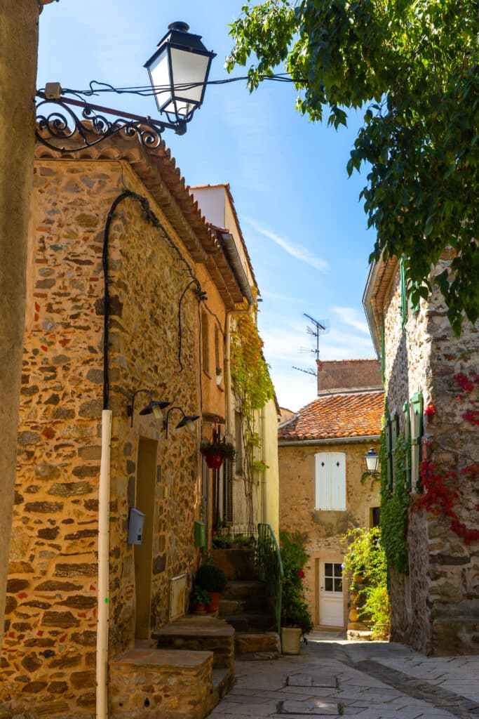Allée étroite en pierre avec bâtiments rustiques, toits en tuiles et végétation luxuriante. Un lampadaire ancien surplombe le chemin sous un ciel bleu.
