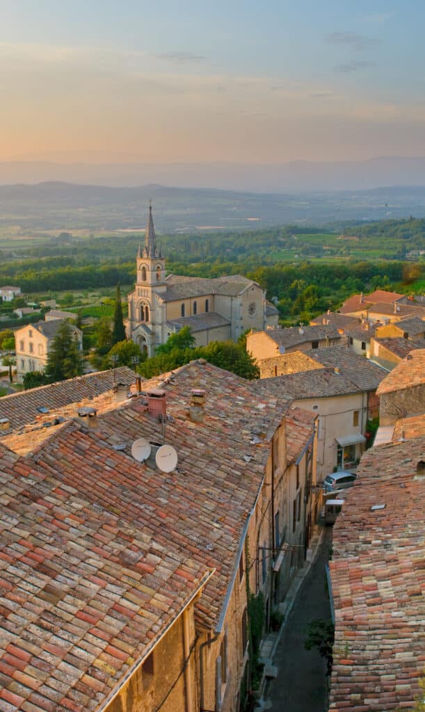 Toits en tuiles d'un village provençal au coucher du soleil, avec une église historique et une vallée verdoyante en arrière-plan.