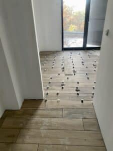 Pose de carrelage imitation bois en cours, avec des croisillons pour l'espacement sur un sol clair, devant une grande baie vitrée.