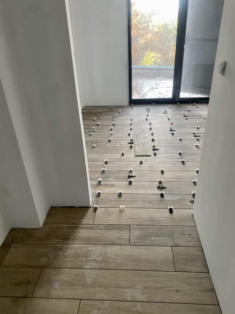 Pose de carrelage imitation bois en cours, avec des croisillons pour l'espacement sur un sol clair, devant une grande baie vitrée.