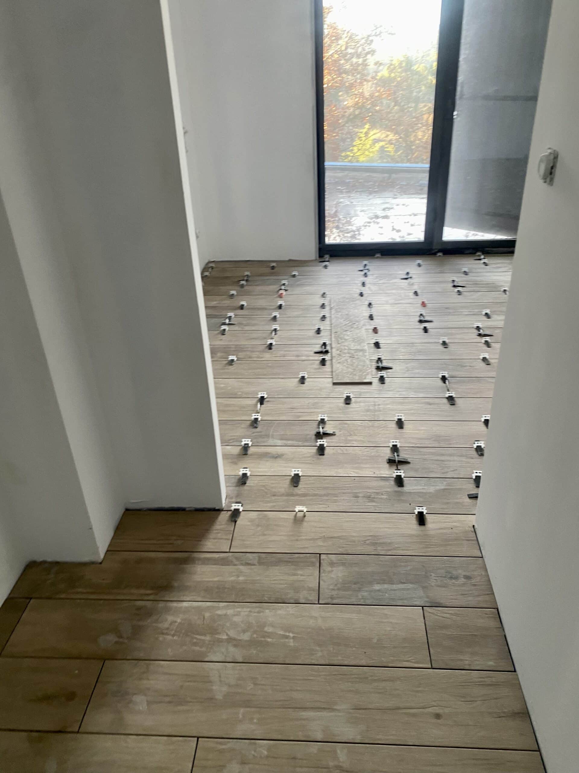 Pose de carrelage imitation bois en cours, avec des croisillons pour l'espacement sur un sol clair, devant une grande baie vitrée.