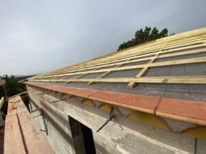 Charpente de toit en construction : liteaux de bois posés sur écran sous-toiture noir avec marquages, au-dessus d'un mur en parpaings et de tuiles de rive.