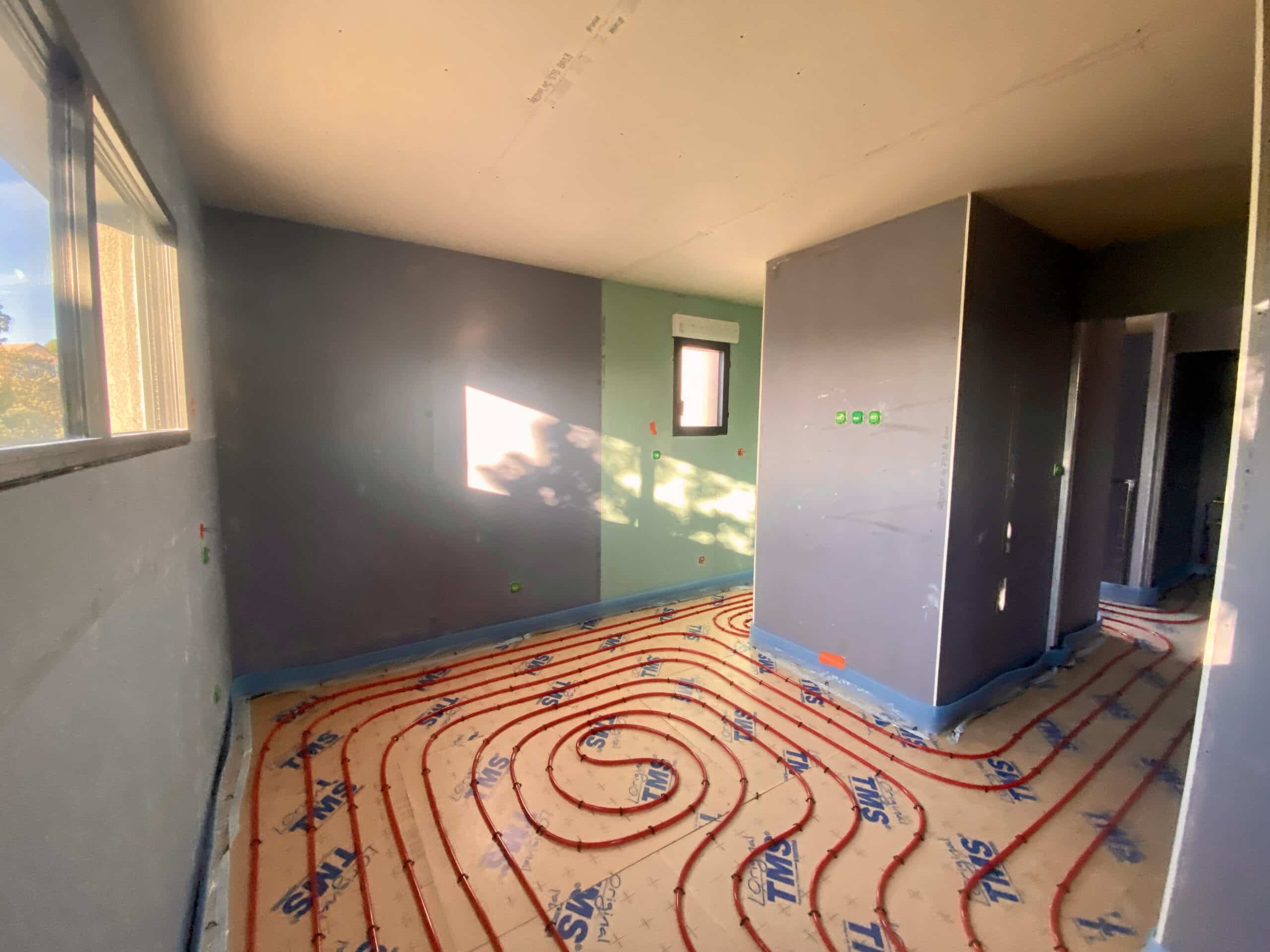 Chantier intérieur : pose de chauffage au sol (tuyaux rouges TMS) sur plaques isolantes avant chape, murs peints gris et vert.