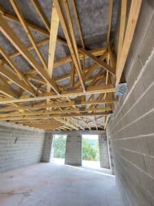 Intérieur d'une maison en construction, murs en parpaings gris, charpente apparente en bois et deux ouvertures donnant sur la verdure.