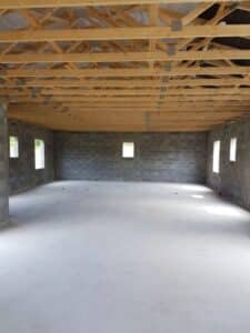 Intérieur brut d'un bâtiment avec murs en parpaings, plancher béton neuf et charpente apparente en bois clair et métal.