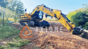 Excavatrice JCB jaune sur chantier de terrassement en terre rouge. Logo AXTOME CONSTRUCTION visible.