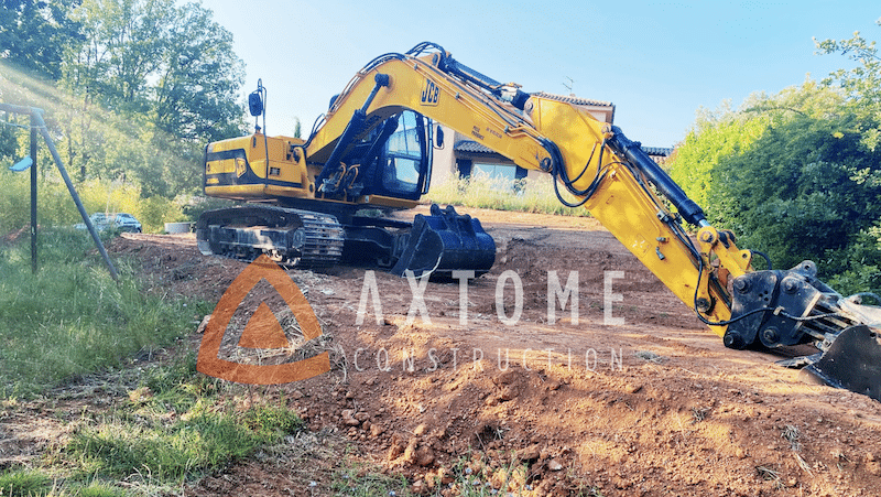 Excavatrice JCB jaune sur chantier de terrassement en terre rouge. Logo AXTOME CONSTRUCTION visible.