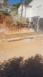 Chantier de fondation en terre rouge, ferraillage et coffrage en bois prêts pour coulage de béton devant une maison aux volets blancs.