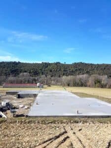 Dalle de béton fraîchement coulée pour une construction, avec des parpaings et une brouette à gauche, sur fond de campagne et de colline boisée.