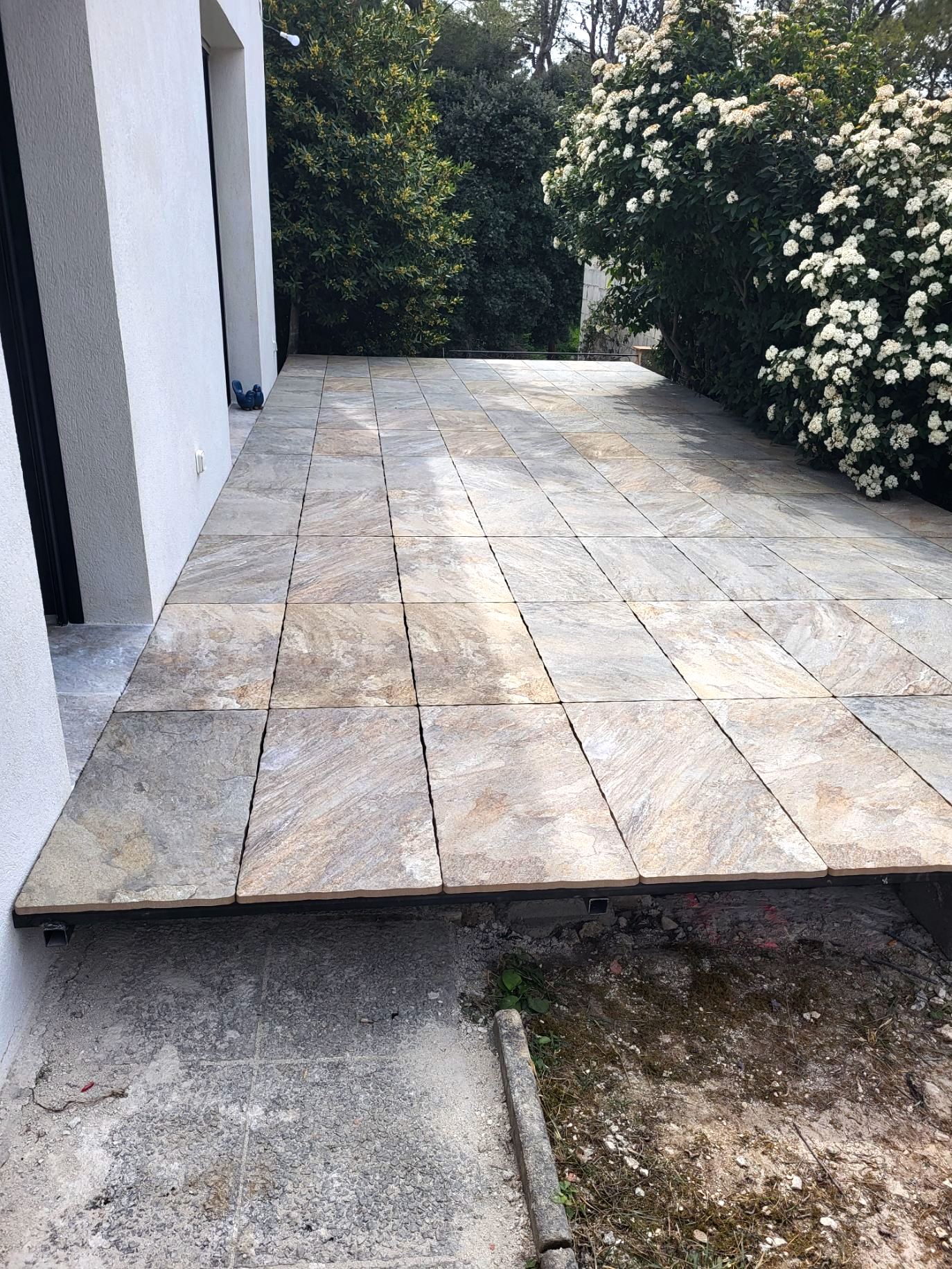 Terrasse en dalles de pierre naturelle multicolore en cours de pose devant une façade blanche et des arbustes fleuris.