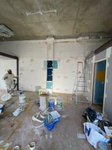 Intérieur de chantier : murs enduits et échantillons de peinture bleue. Plafond brut en béton, ouvrier peintre et matériel de rénovation au sol.