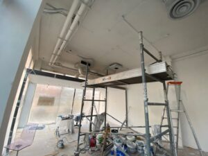 Intérieur en rénovation. Échafaudage, pots de peinture, et échelle sur un chantier. Plafond blanc avec tuyaux apparents et bouche d'aération.