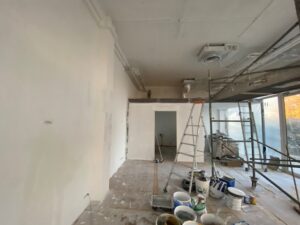 Chantier de rénovation intérieur. Murs apprêtés, échafaudage métallique, échelles, seaux de peinture et conduits de ventilation exposés.