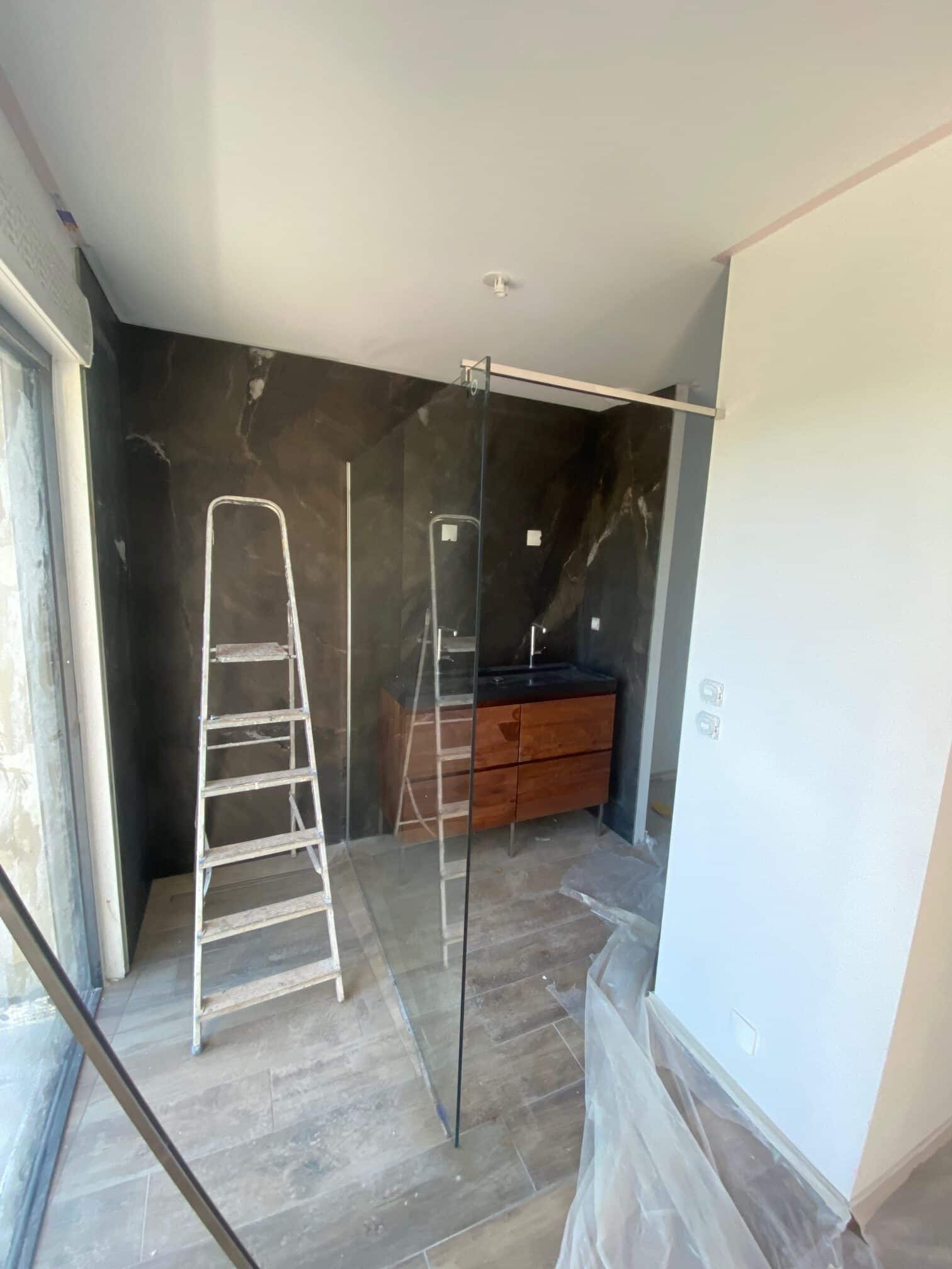 Salle de bain moderne en rénovation : douche italienne, paroi en verre, meuble en bois et mur en pierre sombre.