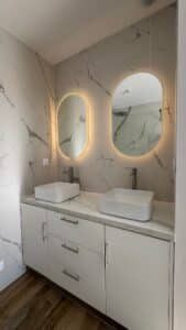 Double vasque sur meuble blanc, murs effet marbre et miroirs ovales rétroéclairés dans une salle de bain moderne.