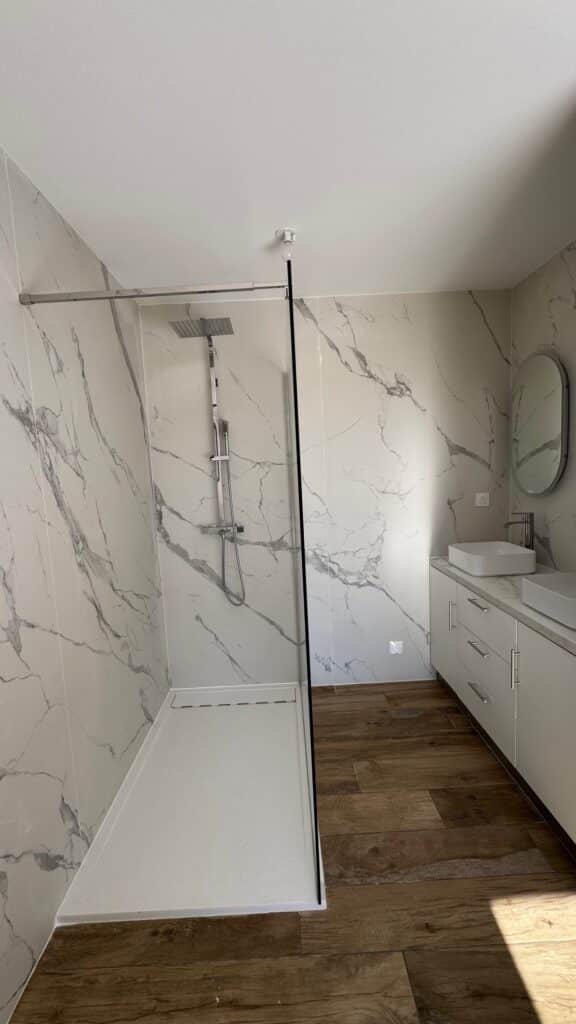Salle de bain moderne avec douche à l'italienne en marbre blanc, double vasque et sol effet bois chaleureux.