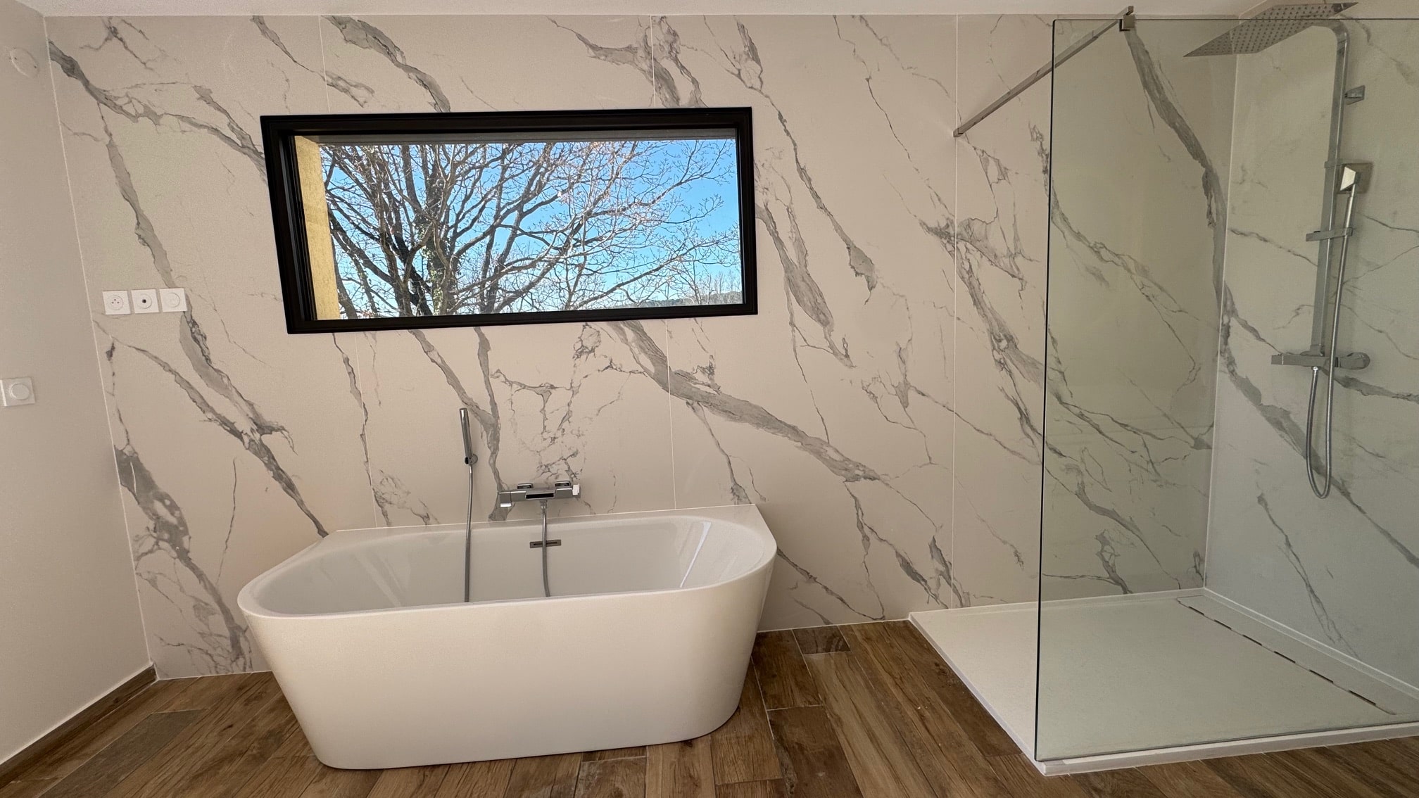 Salle de bain moderne avec baignoire îlot, douche à l'italienne, murs effet marbre et fenêtre sur jardin.