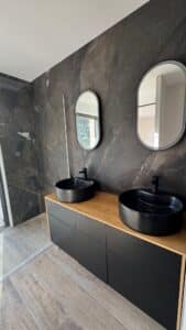 Salle de bain moderne avec double vasque noire, plan de travail en bois et murs effet marbre gris sombre.