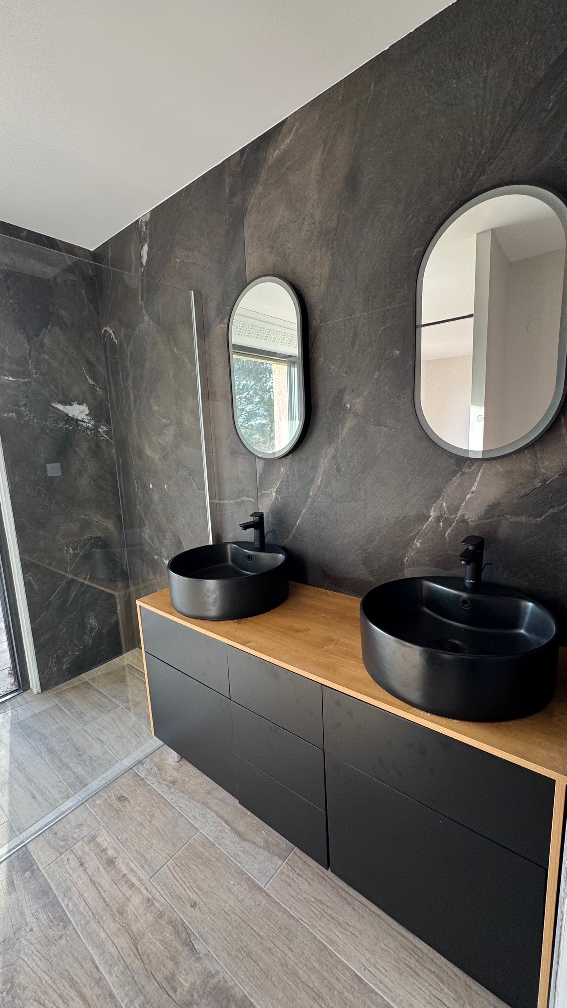 Salle de bain moderne avec double vasque noire, plan de travail en bois et murs effet marbre gris sombre.