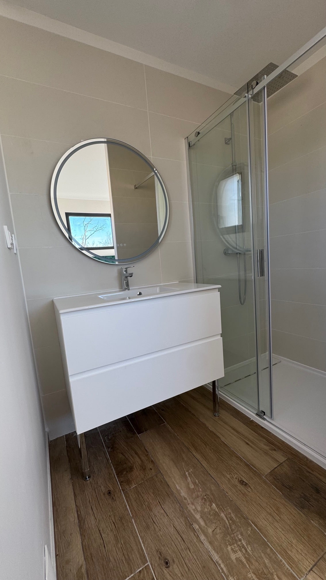 Salle de bain moderne avec meuble vasque blanc, miroir rond LED et douche vitrée sur sol imitation bois.
