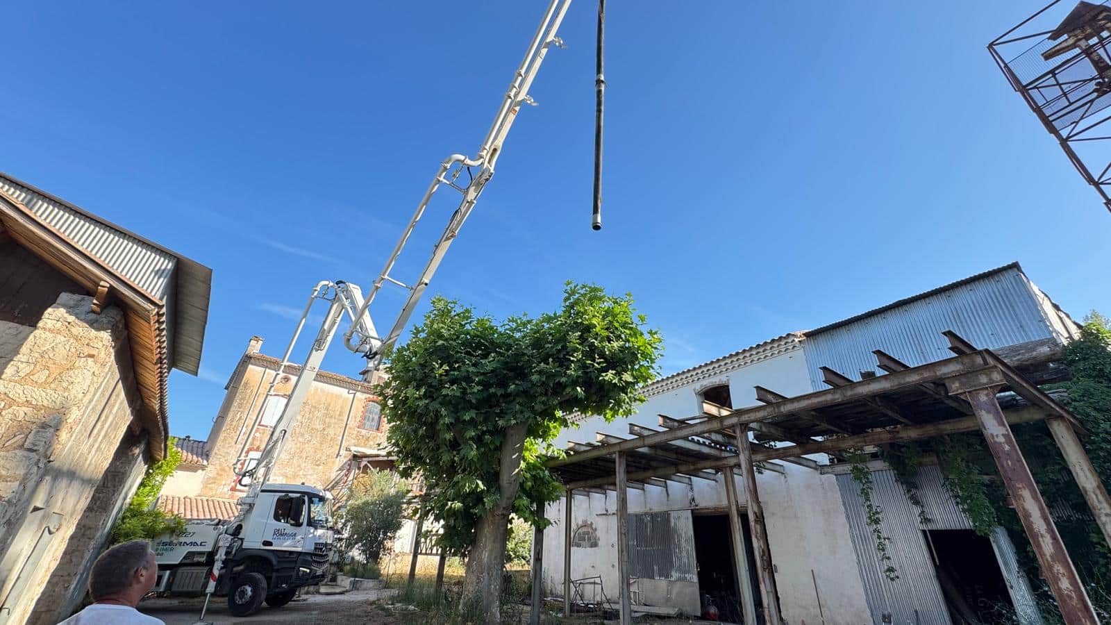 Camion pompe à béton Sermac avec bras articulé au-dessus d'un arbre et d'anciens bâtiments sous ciel bleu.