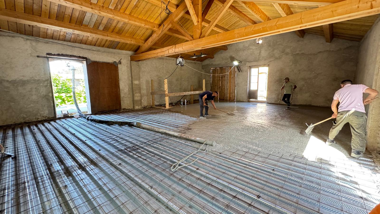 Ouvriers nivelant du béton sur treillis métallique pour une chape de sol dans un bâtiment à charpente bois.